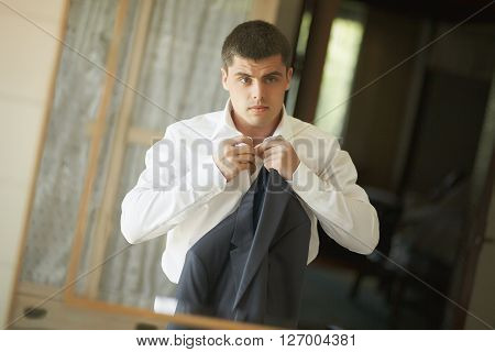 Handsome and strong man putting on official suit. Nice and slender groom wearing wedding suit and putting on cuffs in bedroom looking in mirror.