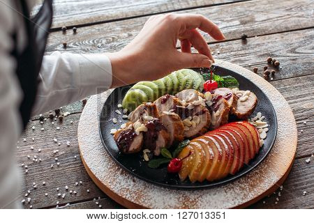 Chief decorated  baked veal on wooden table. Chief decorated  dietary meat with fresh tropical fruits. Top view on food stylist at work. POV on food decoration.
