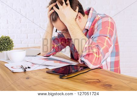 Stressed caucasian male with head in hands over wooden desk with tablet business charts and coffee mug on white brick wall background