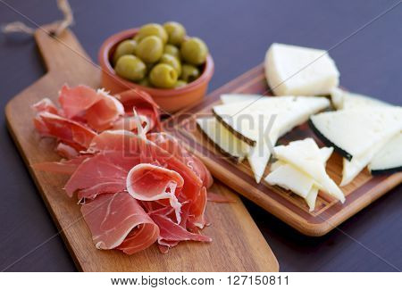 Traditional Spanish Tapas with Sliced Hamon Goat Cheese and Green Olives on Wooden Cutting Board closeup on Dark Wooden background. Focus on Hamon