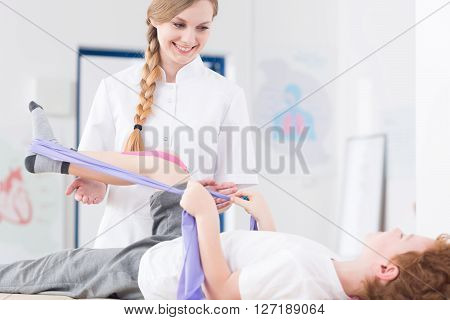 Little patient exercising his leg using elastic bend during physical therapy. Next to him smiling young physiotherapist