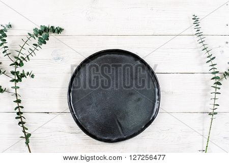 Top view empty black plate on rustic wooden table. Ceramic black plate on white wooden background with free space. Flat lay of handmade black dish on white wooden table.