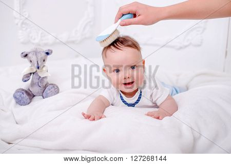 A cute little baby is looking into the camera on the white wall whith blue eyes with bear. Mother combs baby.