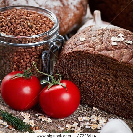 Close up photo of rustic bread on sackcloth, garlic, dill, two tomatoes, oat flakes and jar with buckwheat on planked wooden table