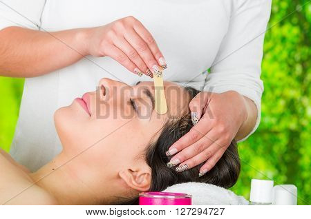 Closeup womans face receiving facial hair waxing treatment, hand using wooden stick to apply wax, beauty and fashion concept.
