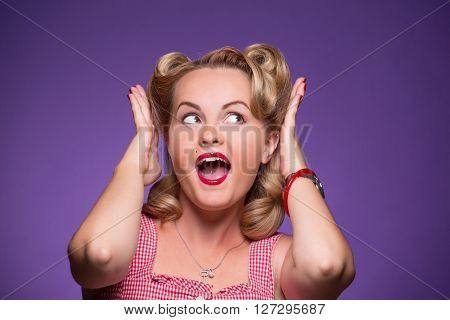 Surprised young sexy pin-up girl screaming isolated on violet background. Astonished and excited girl with red lips looking away with her hands raised.