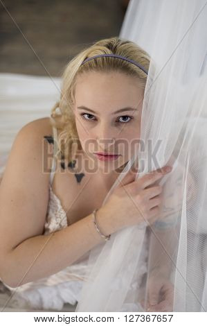 Sexy plus size blonde woman on a bed looking around a lacy curtain.