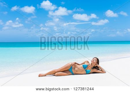 Sexy bikini woman relaxing with tanned slim body on beach vacation paradise getaway. Asian model lying down on perfect white sand sunbathing in tropical Caribbean travel destination. Luxury living.