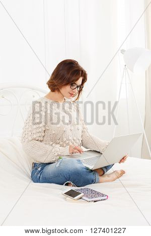Young woman reads from laptop computer in bed. Engrossed Girl in eyeglasses and earphones with mobile phone. Attractive woman in casual clothes at white bed. Internet freelancer. High key, soft tone