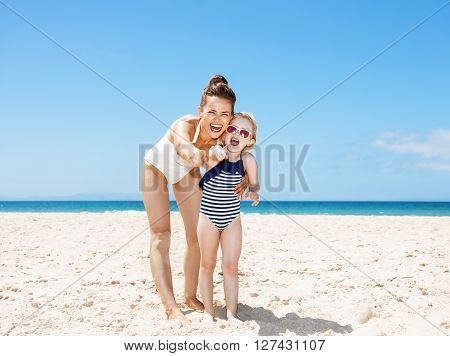 Smiling Mother And Child Pointing In Camera At Sandy Beach