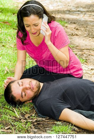 Young man lying down with medical emergency, woman sitting by his side calling for help, outdoors environment.