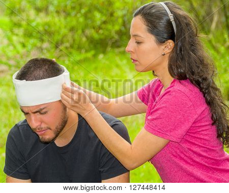 Young man with head injury receiving treatment and bandage around skull from woman, outdoors environment.