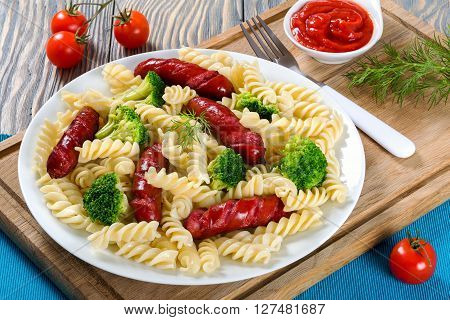 Delicious Spiral Pasta salad with broccoli and grilled sausages decorated with dill on a white dish with tomato sauce in a gravy boat and fork on a cutting board studio lights close-up