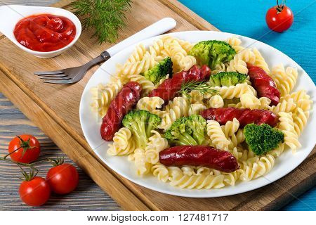 Delicious Spiral Pasta salad with broccoli and grilled sausages decorated with dill on a white dish with tomato sauce in a gravy boat and fork on a cutting board studio lights top view