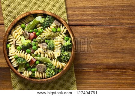 Pasta primavera with green asparagus pea broccoli mushroom and tomato in cream sauce served in wooden bowl photographed overhead on dark wood with natural light