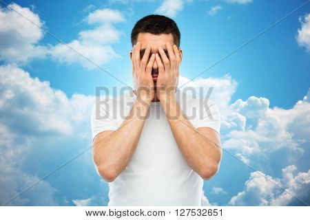 people, crisis, emotions and stress concept - man in white t-shirt covering his face with hands over blue sky and clouds background