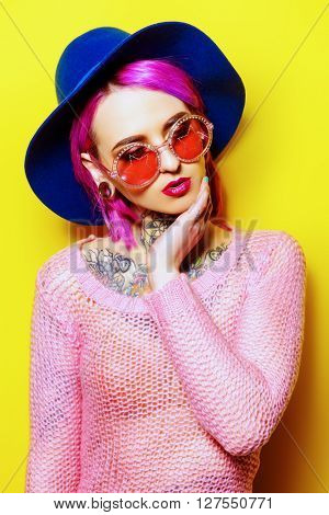 Pretty girl with crimson hair wearing bright clothes and sunglasses posing over yellow background. Bright style, fashion. Optics style. Tattoo.