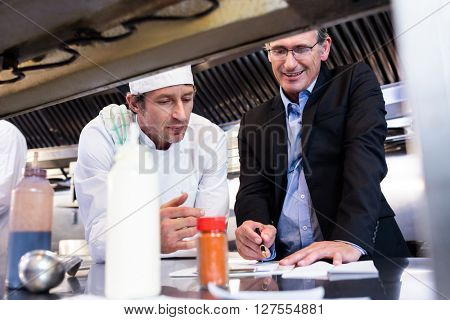 Male restaurant manager writing on clipboard while interacting to head chef in commercial kitchen