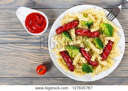 Delicious Spiral Pasta salad with broccoli and grilled sausages decorated with dill on a white dish with tomato sauce in a gravy boat studio lights top view