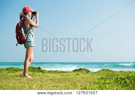 Young lady with rucksack taking a picture of the sea standing on the green glage