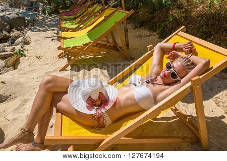 Beautiful  woman in white bikini, sunglasses and hat sunbathing on a deck chair on Paradise Beach, the most glamorous beach of Patong, Phuket in Thailand.