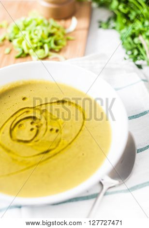 A close up photograph of a white bowl with classic vegetarian potato and leek soup decorated with olive oil. Green parsley, wood peppermill and sliced leek on background. White rustic wood background