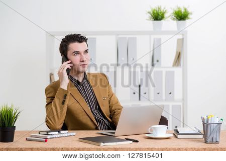 Thoughtful caucasian businessman in brown jacket sitting at office desk with laptop and other items and having a telephone conversation