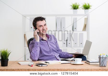 Smiling caucasian businessperson in striped purple shirt sitting at office desk with laptop and other items and having a telephone conversation