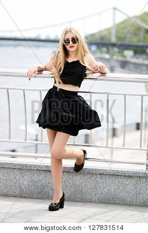 Full length outdoor portrait of a beautiful young blonde in bkack skirt blouse sunglasses and high-heeled shoes on a light background