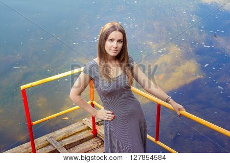 Beautiful young woman with long hair in striped casual dress standing on the old wooden pier above the silty river water