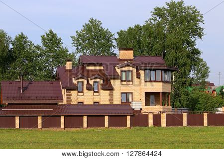 Bright yellow cottage among green trees and brown fence at summer day