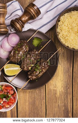 Grilled kebabs with vegetable salad, couscous and seasonings over wooden table