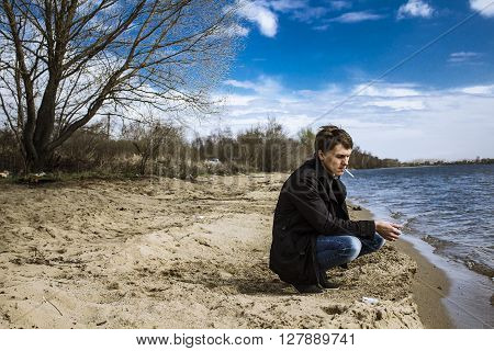 The man on the beach in the thoughts of his beloved, with a cigarette, trenchcoat, sand and sun, a leafless tree