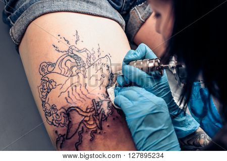 Top view of the process of tattooing in a professional salon. Tattoo design Shark punched harpoon. Tattoos art on human body skin - clients leg.