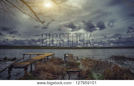 Old wooden pier with dry reed on sunset. Cloudy weather. Dramatic sky