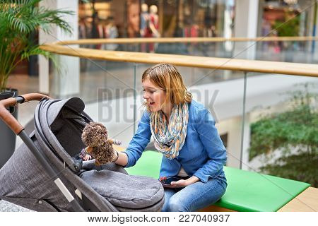Young Smiling Woman Sitting On Bench With Stoller In Shopping Center And Showing Soft Toy To Her Bab