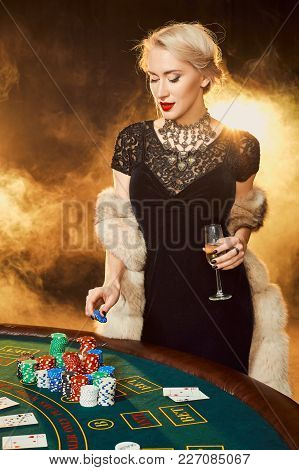 Young Woman In Evening Dress With Chips In Hand Standing Near Poker Table With Glass Of Champagne