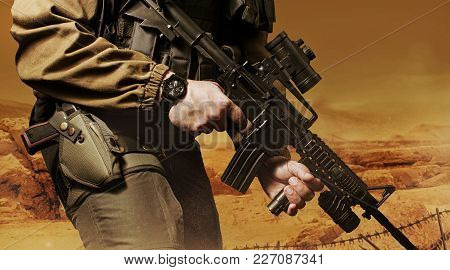 Photo of a soldier in military outfit holding a gun and bulletproof vest on orange desert background.