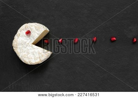 Camembert Or Brie Cheese Cut Out Like Pacman Eating Pomegranate Seeds On Black Slate Background. Fun