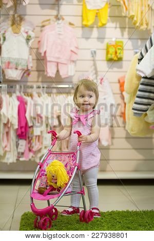 Little Blonde Girl Smiling And Clapping Her Hands In The Kids' Store With The Toy Baby Carriage