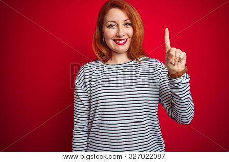 Young redhead woman wearing strapes navy shirt standing over red isolated background showing and pointing up with finger number one while smiling confident and happy.