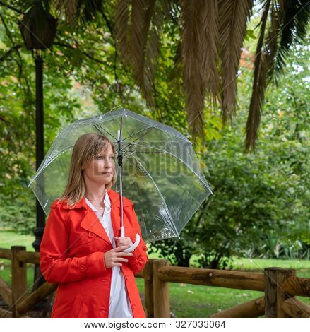 Caucasian Girl With Red Gabardine Walks Through A Park Under A Transparent Umbrella.