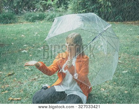 Caucasian Girl With Red Gabardine Walks Through A Park Under A Transparent Umbrella.