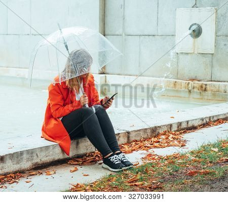 Caucasian Girl With Red Gabardine Walks Through A Park Under A Transparent Umbrella.