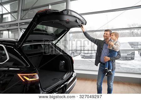 Father Holding Little Son On Hands And Looking At Big Capacious Trunk Of Automobile In Auto Salon. C