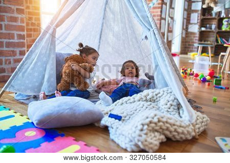 Adorable toddlers lying down over blanket inside tipi smiling and playing with doll at kindergarten