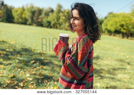 Smiling Beautiful Young Woman Wearing Sweater And Holding Disposable Cup Of Coffee In The City Park.
