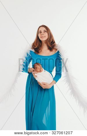 A Girl In A Blue Dress With Angel Wings Holding A Newborn Baby On A White Background. An Angel-woman