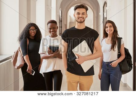 Multiracial Group Of Four Smiling Students Dressed Casual Standing In University Hall, Looking At Ca