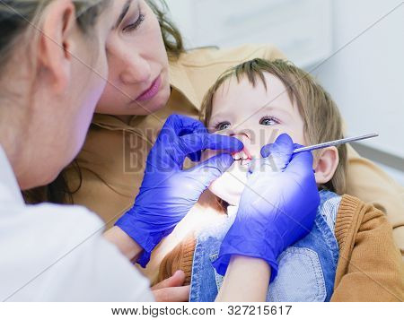 The Doctor Looks At The Teeth Of A Small Child. The Baby Is Sitting In The Arms Of Mom. Dentist Exam